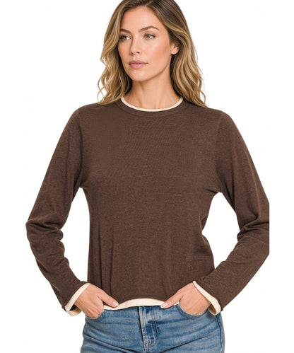 A woman wearing a brown long-sleeve knit top with contrasting cream trim at the crew neck and cuffs, paired with blue jeans, against a plain white background.