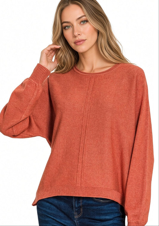 A woman wearing a rust-colored front seam knit sweater with balloon sleeves and a crew neck, paired with dark blue jeans, against a plain white background.