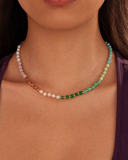 A close-up of a model wearing a gold necklace made of small, faceted crystals beads that transition through peach, white, green, and light blue, styled with a dark purple top.