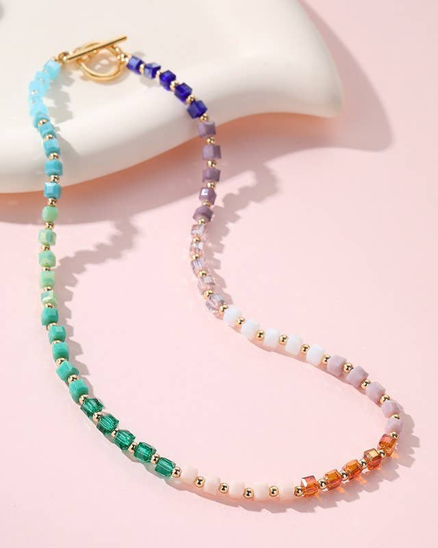 A beaded necklace with a gold clasp, featuring a rainbow gradient of faceted beads in blue, green, purple, pink, and orange against a soft pink background.