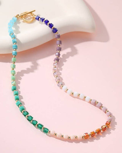 A beaded necklace with a gold clasp, featuring a rainbow gradient of faceted beads in blue, green, purple, pink, and orange against a soft pink background.