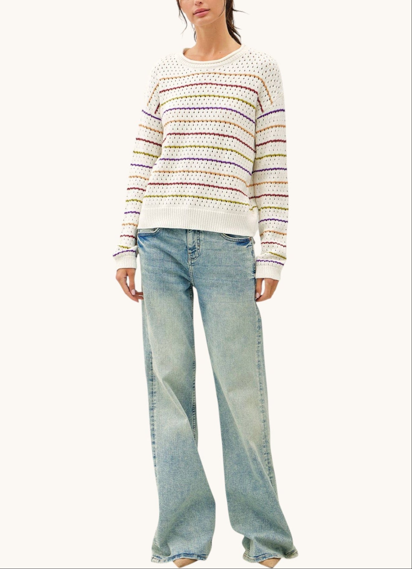 A woman modeling a cream knit sweater with thin multicolored horizontal stripes and light-wash wide-leg jeans against a plain off-white background.