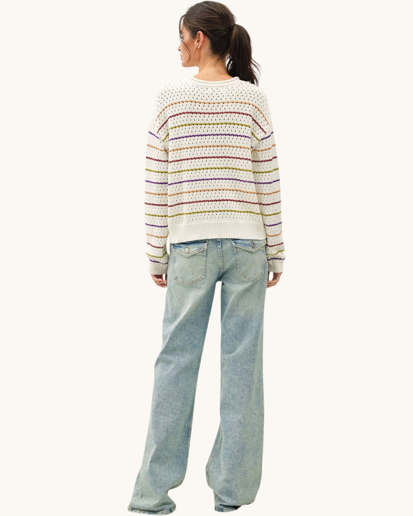A woman shown from the back wearing a cream knit sweater with horizontal multicolor stripes and light-wash wide-leg jeans against a plain off-white background.