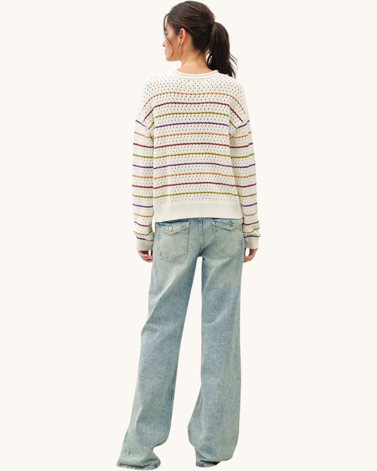 A woman shown from the back wearing a cream knit sweater with horizontal multicolor stripes and light-wash wide-leg jeans against a plain off-white background.