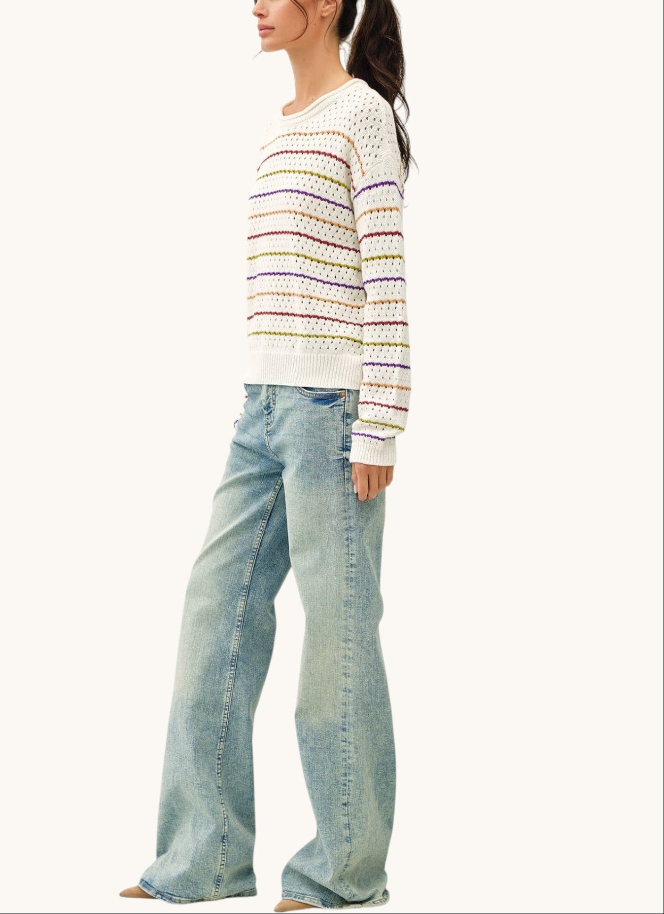 A woman modeling a white knit sweater with thin multicolored horizontal stripes and light-wash wide-leg jeans against a plain off-white studio background.