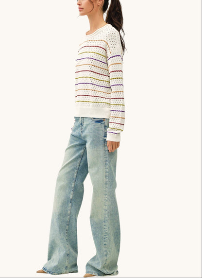 A woman modeling a white knit sweater with thin multicolored horizontal stripes and light-wash wide-leg jeans against a plain off-white studio background.