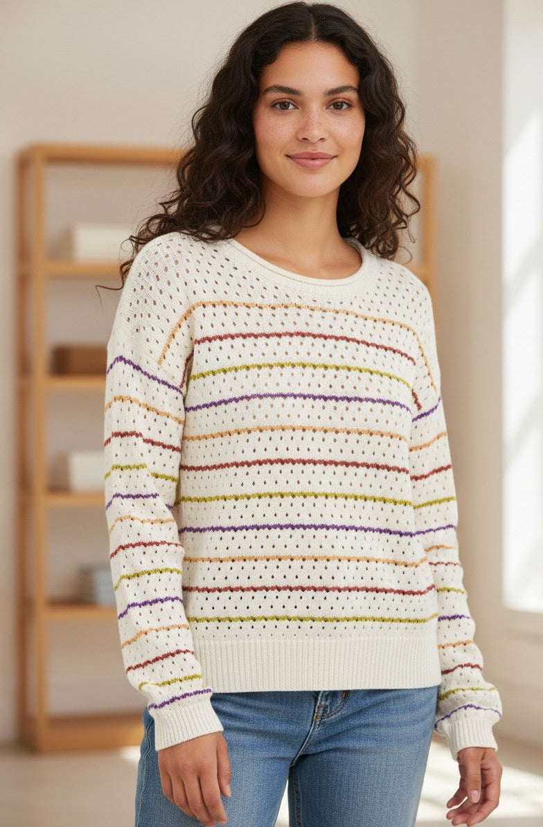 A woman wearing a cream-colored knit sweater with horizontal multicolor stripes in maroon, orange, green and purple, paired with blue jeans, standing in a brightly lit room.