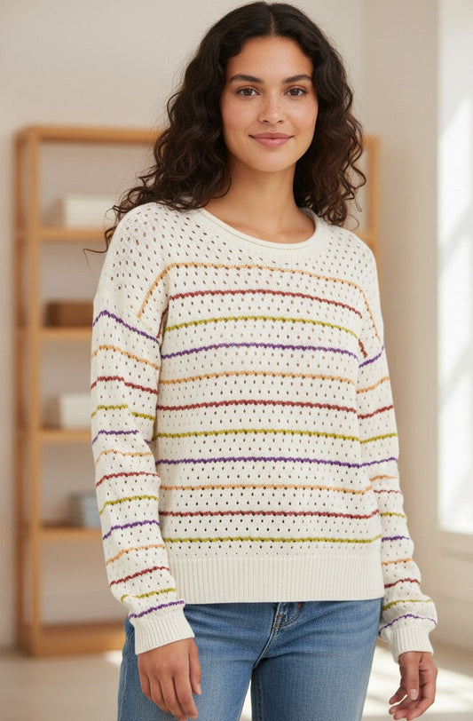 A woman wearing a cream-colored knit sweater with horizontal multicolor stripes in maroon, orange, green and purple, paired with blue jeans, standing in a brightly lit room.