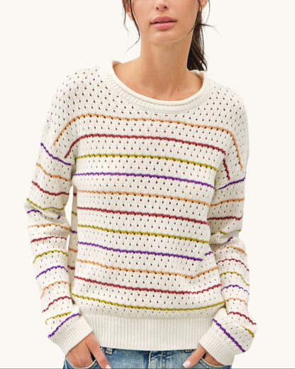 A woman wearing a cream-colored knit sweater with horizontal multicolor stripes in red, orange, yellow, green, and purple, paired with blue jeans against a plain light background.