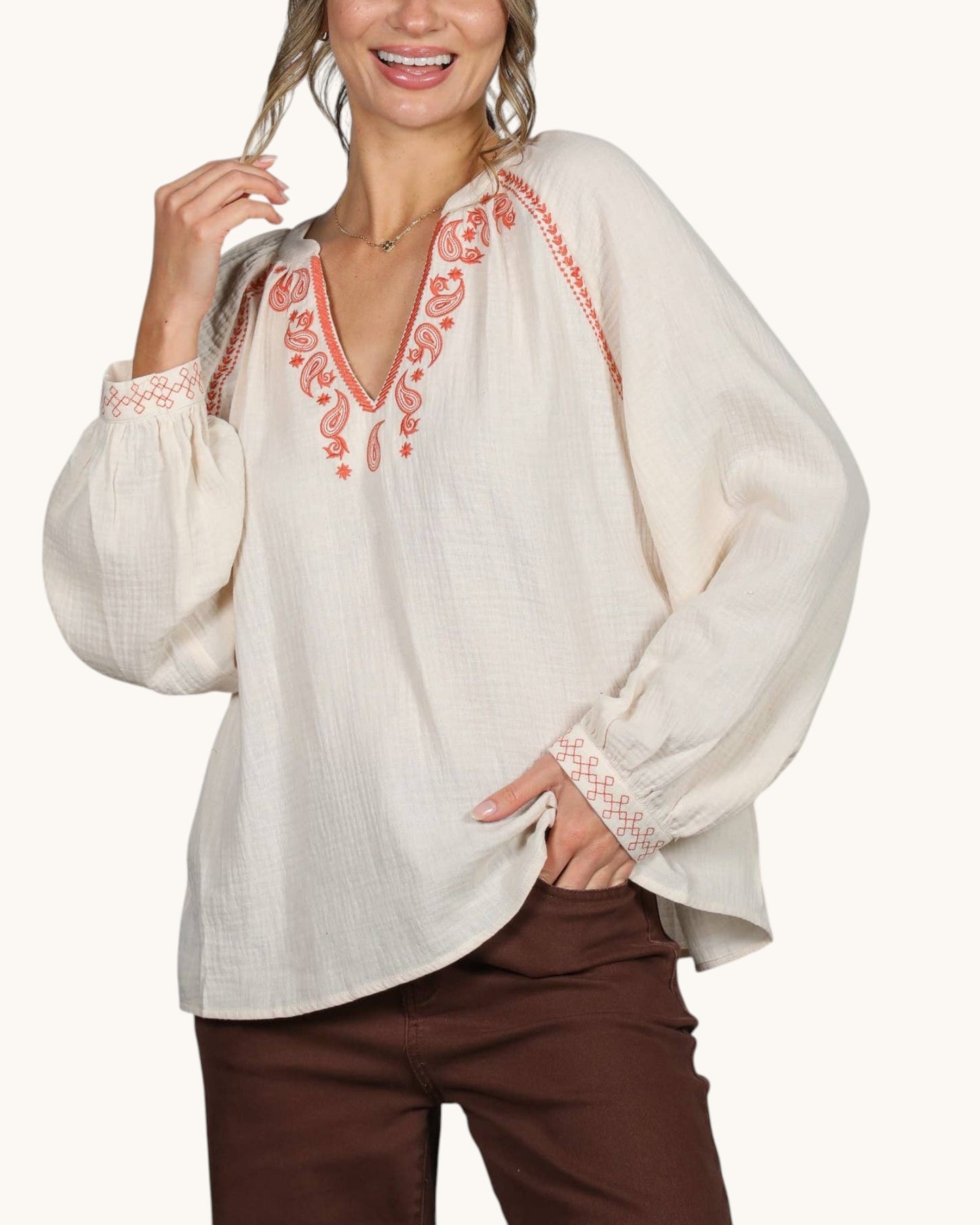 A woman wearing a loose off-white blouse with coral-orange embroidered detailing around the V-neck and cuffs, paired with dark brown pants, against a plain light background.