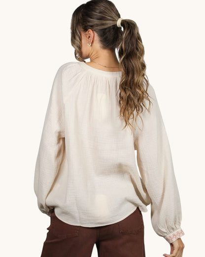 A woman shown from the back wearing a cream-colored, textured long-sleeve blouse with gathered cuffs and brown pants, set against a plain off-white background.