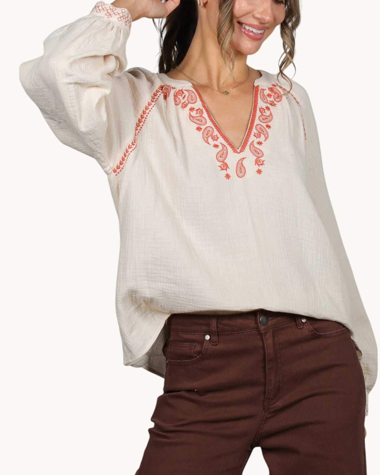 A woman wearing a loose off-white blouse with coral-red embroidered detailing around the V-neck and on the sleeve cuffs, paired with brown pants, against a plain light background.