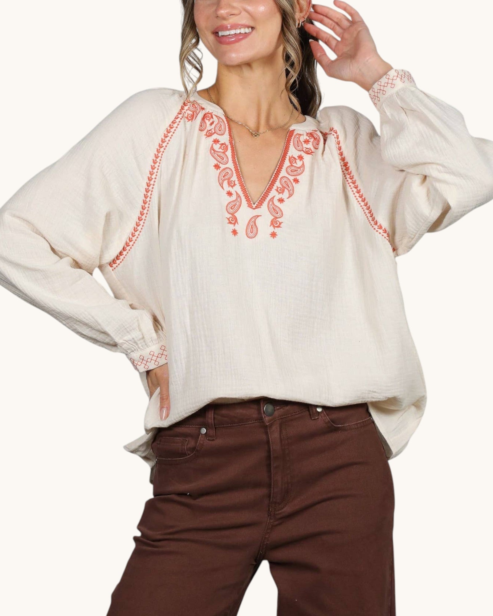 A woman wearing a cream-colored long-sleeve blouse with coral-red paisley embroidery around the V-neck and cuffs, paired with brown pants, against a plain light background.