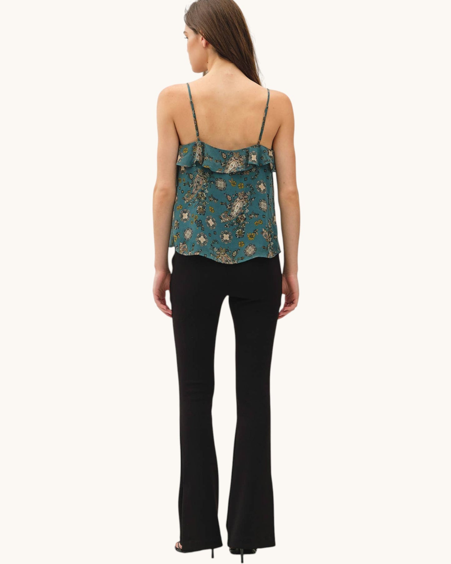 A woman shown from the back wearing a teal floral spaghetti-strap camisole top and black flared pants against a plain off-white background.