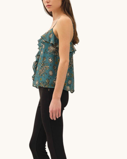 A woman shown in side profile wearing a teal ruffled sleeveless top with a floral print and black flared pants against a plain light background.