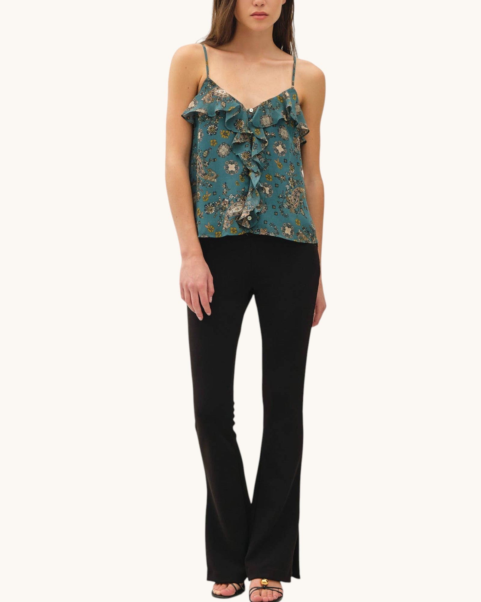 A model wearing a teal floral ruffled camisole top with black flared pants, shown against a plain off-white background.