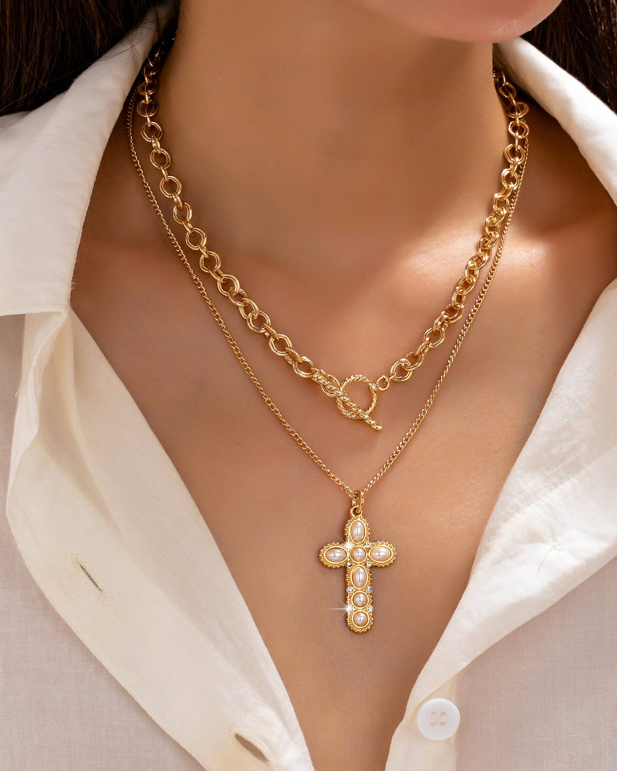 A close-up of a model wearing a double-layered gold necklace. The top layer features a chunky chain with a toggle clasp, and the bottom layer chain has a statement cross pendant encrusted with small white pearls, worn over a white collared shirt.