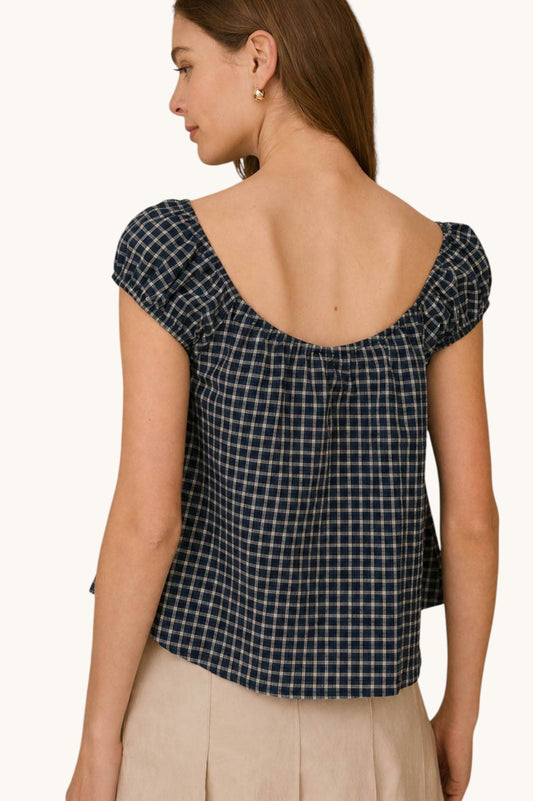 A woman shown from the back wearing a navy and beige plaid top with cap sleeves and a wide scoop neckline, paired with light beige bottoms, against a plain off-white background.