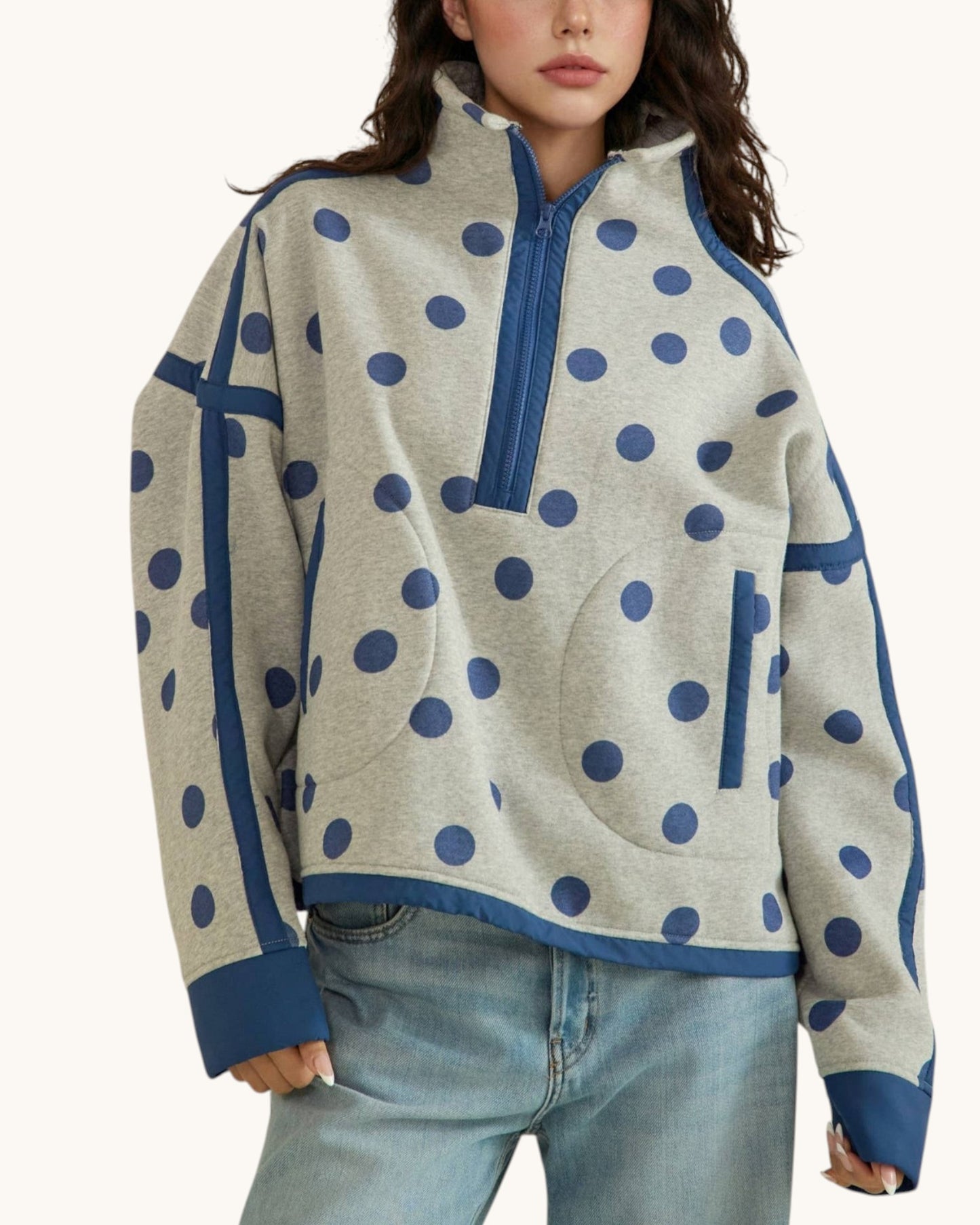 A woman wearing a light heather gray quarter-zip pullover sweater with navy blue polka dots and solid navy trim, paired with light-wash blue jeans, against a plain off-white background.