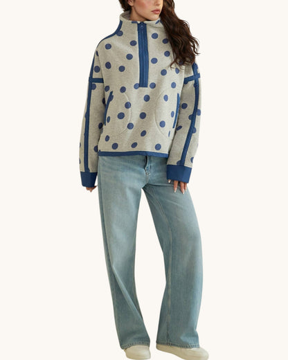 A model wearing a light gray heathered quarter-zip fleece pullover with blue polka dots and blue trim, paired with light-wash wide-leg jeans and white sneakers, against a solid off-white background.