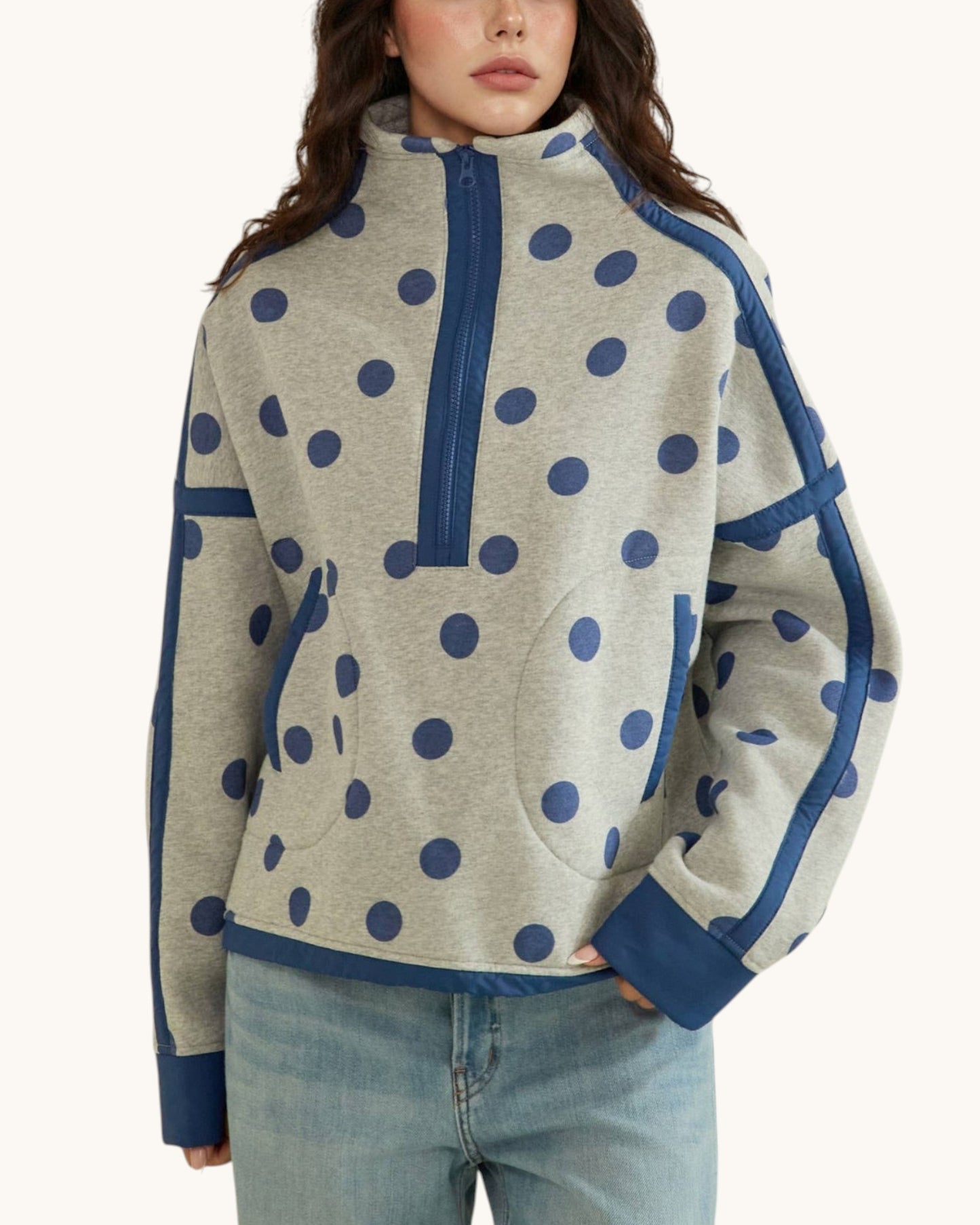 A woman wearing a heather gray quarter-zip pullover sweatshirt with navy blue polka dots and solid navy blue trim, paired with light-wash blue jeans against a plain off-white background.