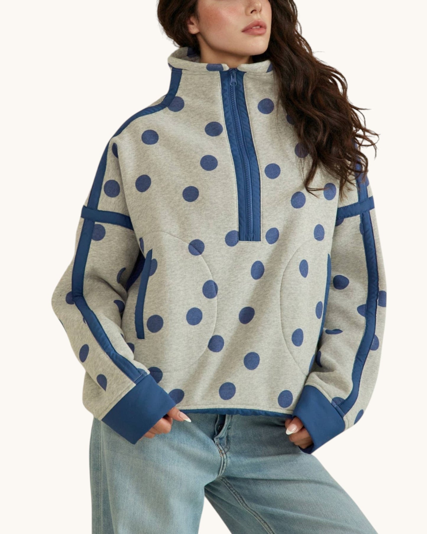 A woman wearing a light heather gray half-zip fleece pullover with navy blue polka dots and solid navy trim, paired with light-wash blue jeans, against a plain off-white background.