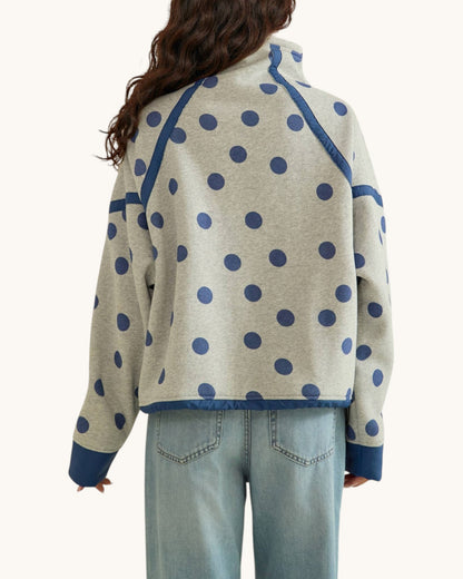 Back view of a woman wearing a light heather gray sweatshirt with navy blue polka dots and solid navy blue trim, paired with light-wash blue jeans against a plain off-white background.