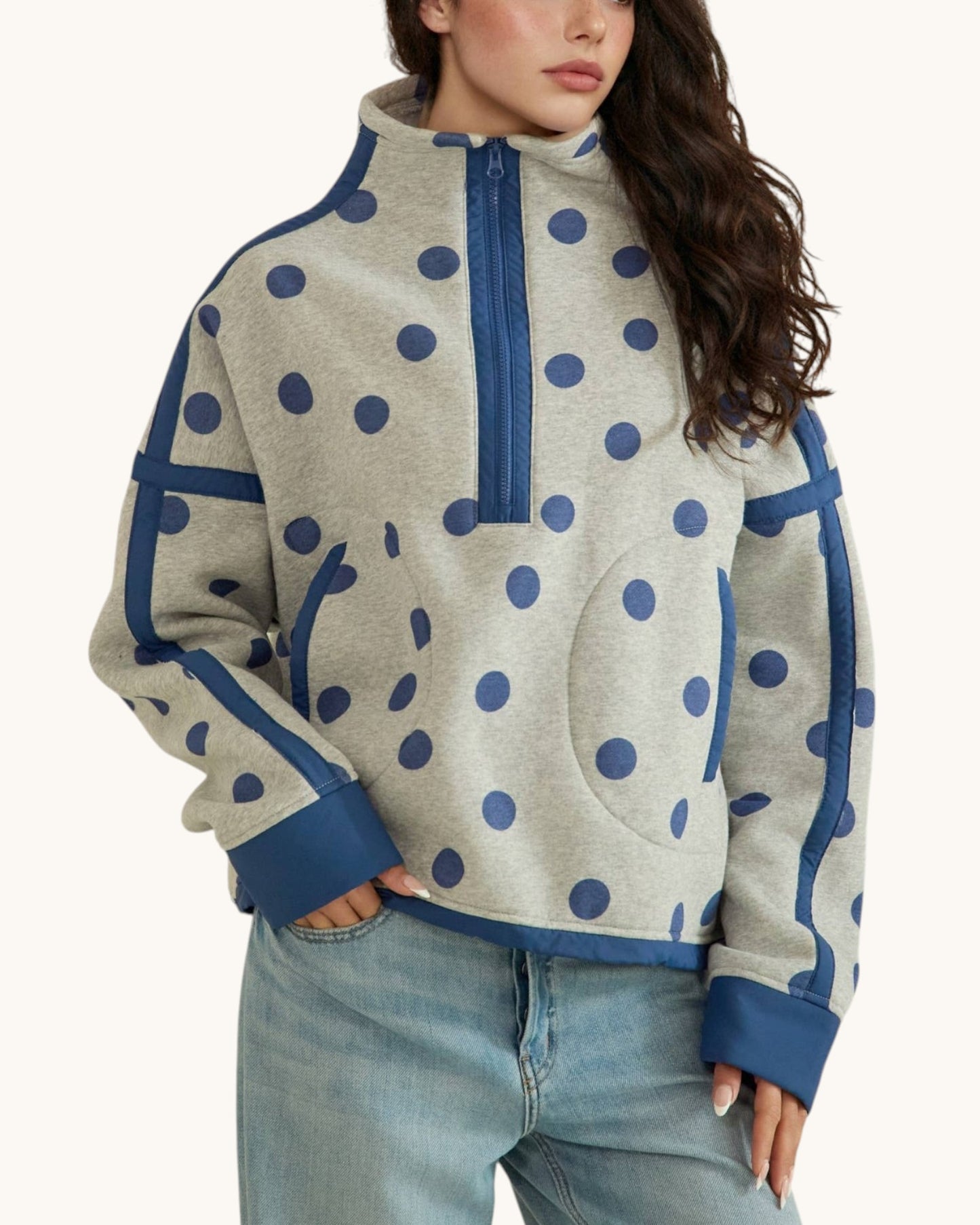 A woman wearing a light heather gray quarter-zip fleece pullover with navy blue polka dots and solid navy trim, paired with light-wash blue jeans against a plain off-white background.