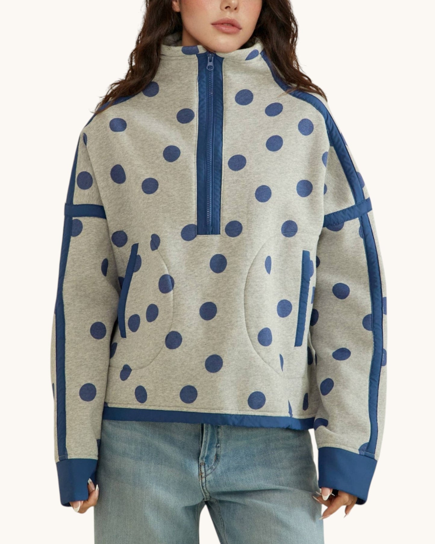 A woman wearing a light heather gray half-zip sweatshirt with navy blue polka dots and solid navy blue trim, paired with light-wash blue jeans against a plain off-white background.