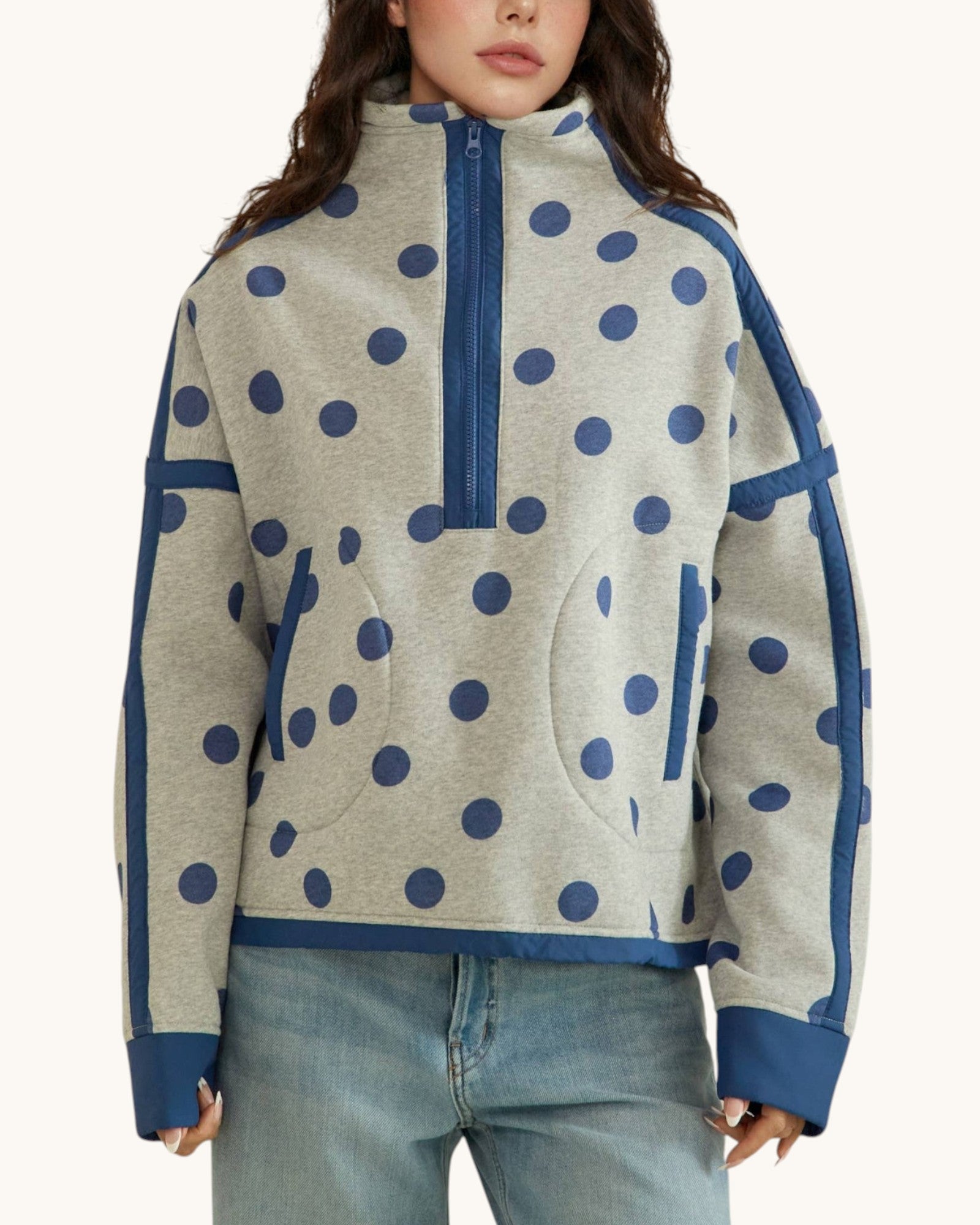 A woman wearing a light heather gray half-zip sweatshirt with navy blue polka dots and solid navy blue trim, paired with light-wash blue jeans against a plain off-white background.