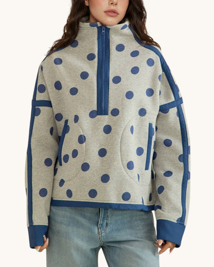 A woman wearing a light heather gray half-zip sweatshirt with navy blue polka dots and solid navy blue trim, paired with light-wash blue jeans against a plain off-white background.
