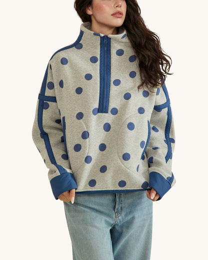 A woman wearing a light heather gray quarter-zip fleece pullover with large navy blue polka dots and solid navy blue trim on the collar, cuffs, and side panels, paired with light-wash blue jeans against a plain off-white background.