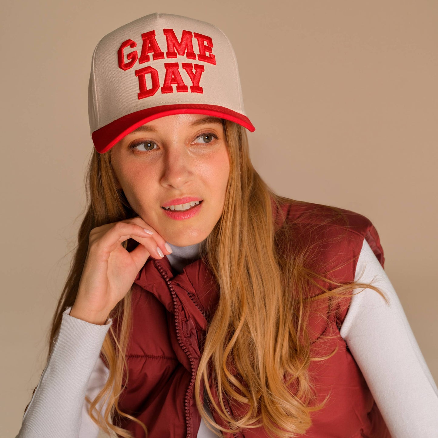 Woman wearing a 'GAME DAY' cap with a beige and red design, standing against a beige background.