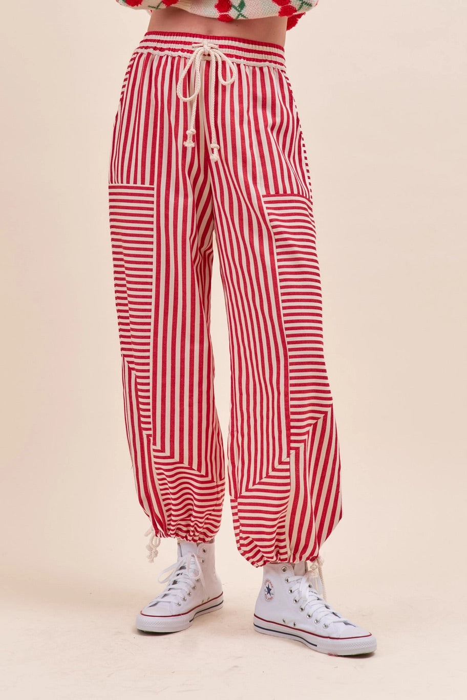 Red and white vertically striped harem style parachute pants with a drawstring waist and elastic cuffs, shown on a model wearing white high-top sneakers.