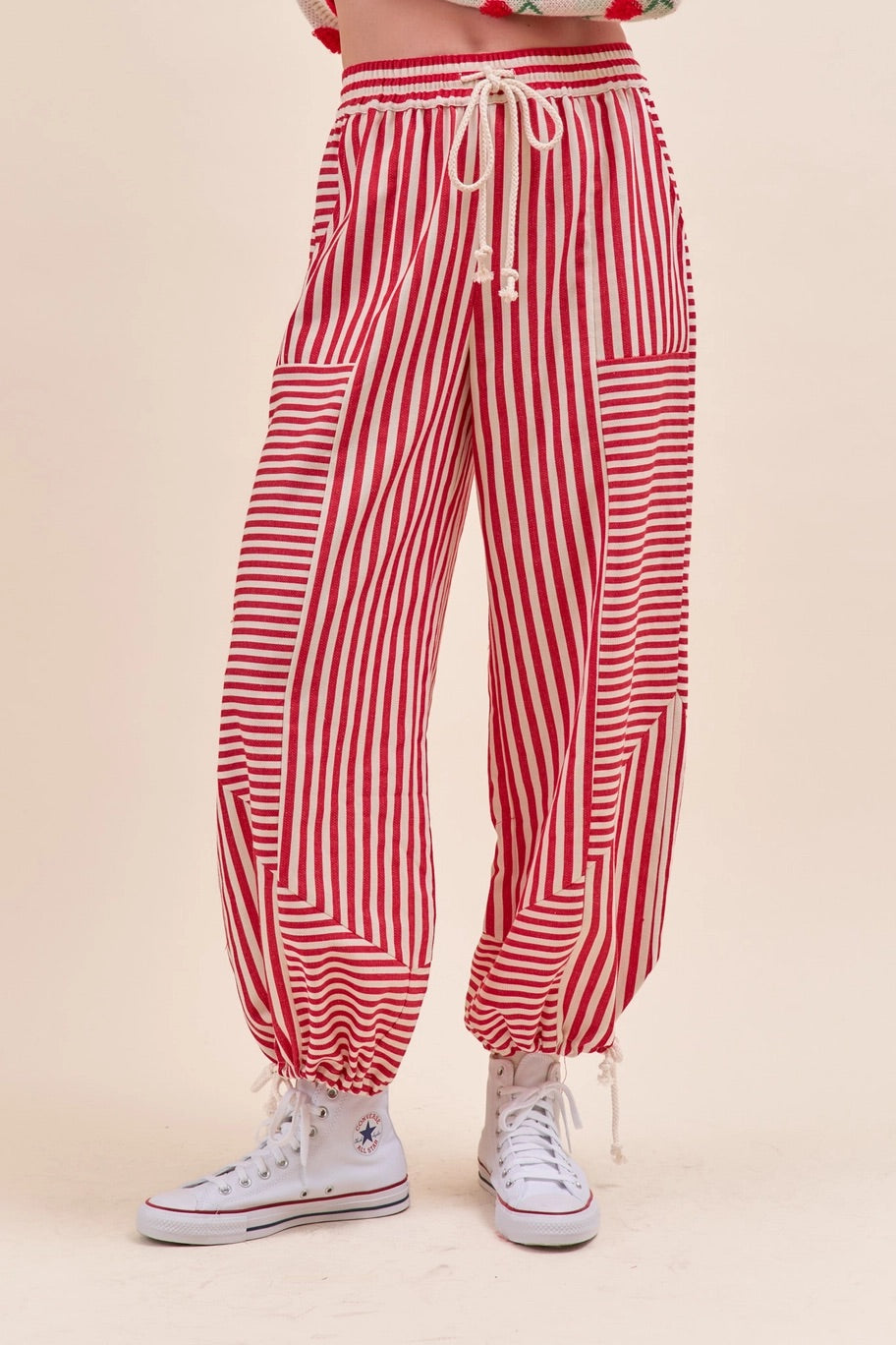Red and white vertically striped harem-style pants with a cream drawstring waist, side pockets, and elastic gathered cuffs, shown on a model wearing white high-top sneakers.