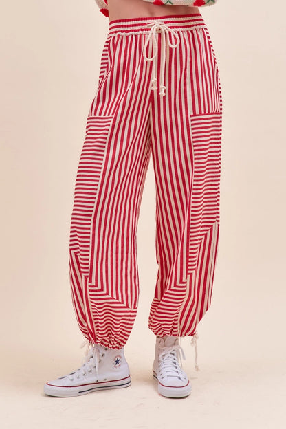 Red and white vertically striped harem-style pants with a cream drawstring waist, side pockets, and elastic gathered cuffs, shown on a model wearing white high-top sneakers.