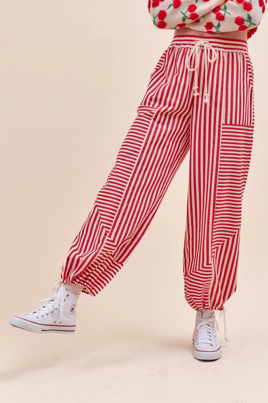 Red and white vertically striped parachute pants with a drawstring waist and side pockets, shown on a model wearing white sneakers against a solid beige background.