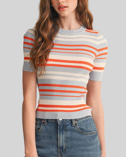 A woman wearing a short-sleeve ribbed knit top with horizontal coral/orange, cream, and light gray-blue stripes, paired with blue jeans, against a solid gray background.