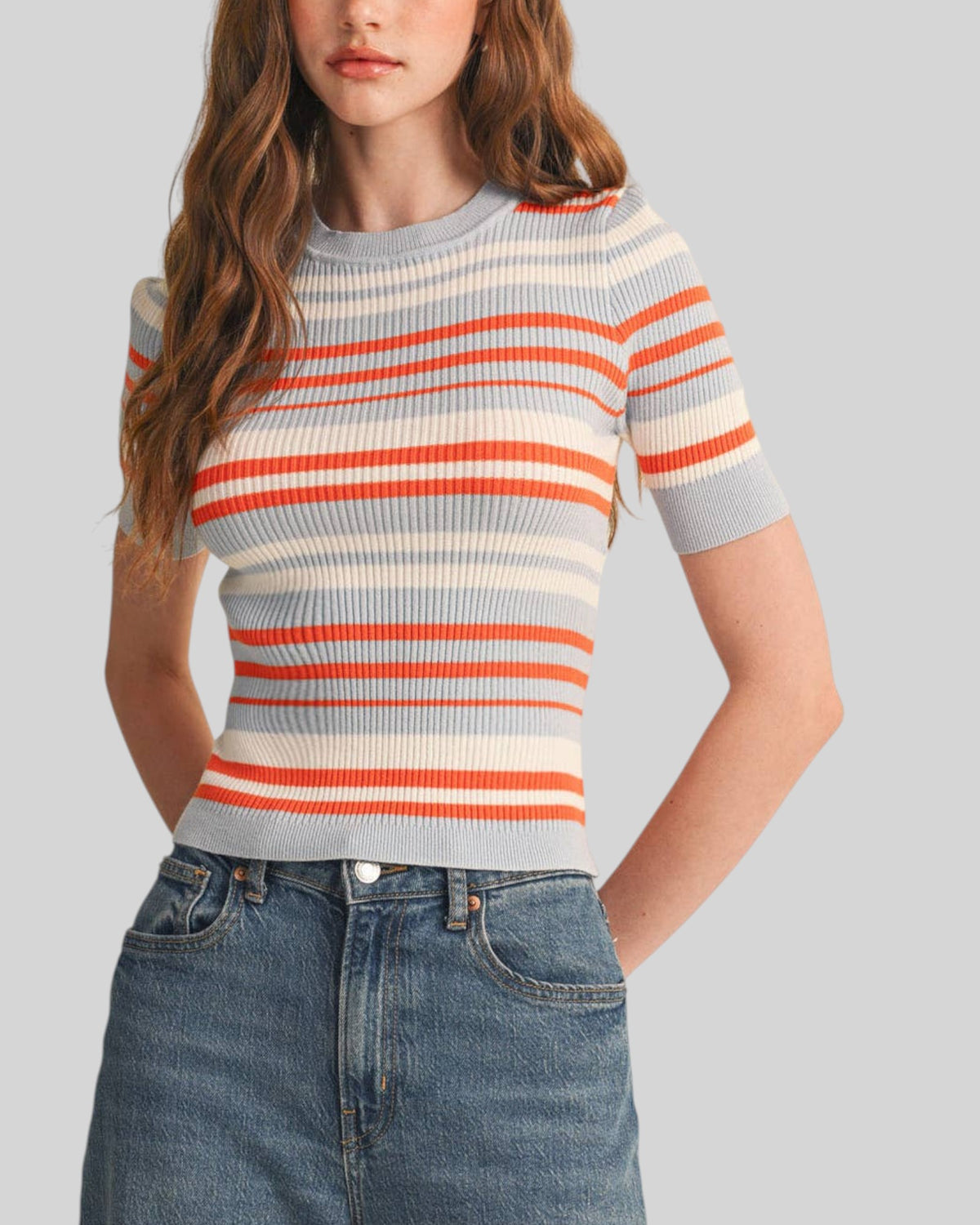 A woman wearing a short-sleeve ribbed knit top with horizontal stripes in orange, cream, and light gray-blue, paired with blue jeans, against a solid light gray background.