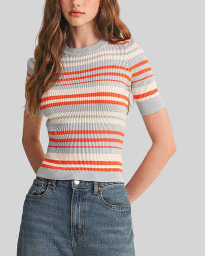A woman wearing a short-sleeve ribbed knit top with horizontal stripes in orange, cream, and light gray-blue, paired with blue jeans, against a solid light gray background.