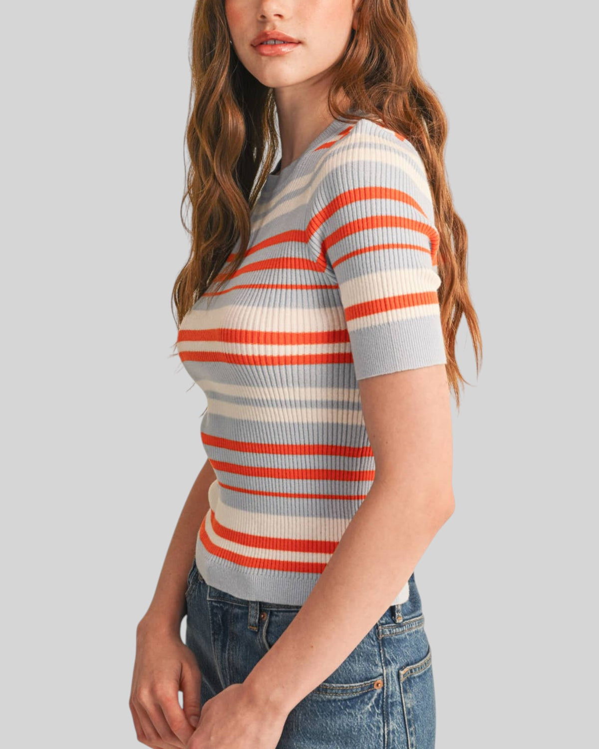 A woman wearing a short-sleeve ribbed knit top with horizontal stripes in light gray/blue, off-white, and orange, paired with blue jeans, shown against a solid light gray background.