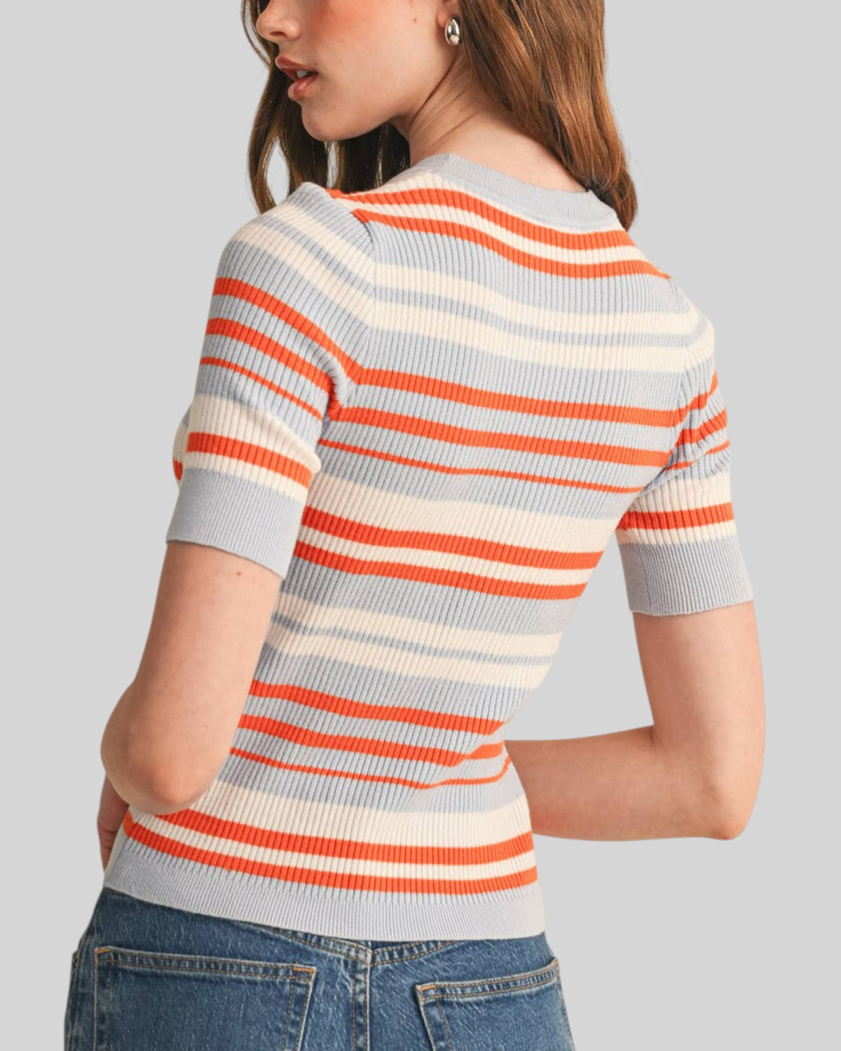 A woman wearing a short-sleeve ribbed knit sweater with horizontal orange, cream, and light gray -blue stripes, shown from a three-quarters back view against a solid gray background.