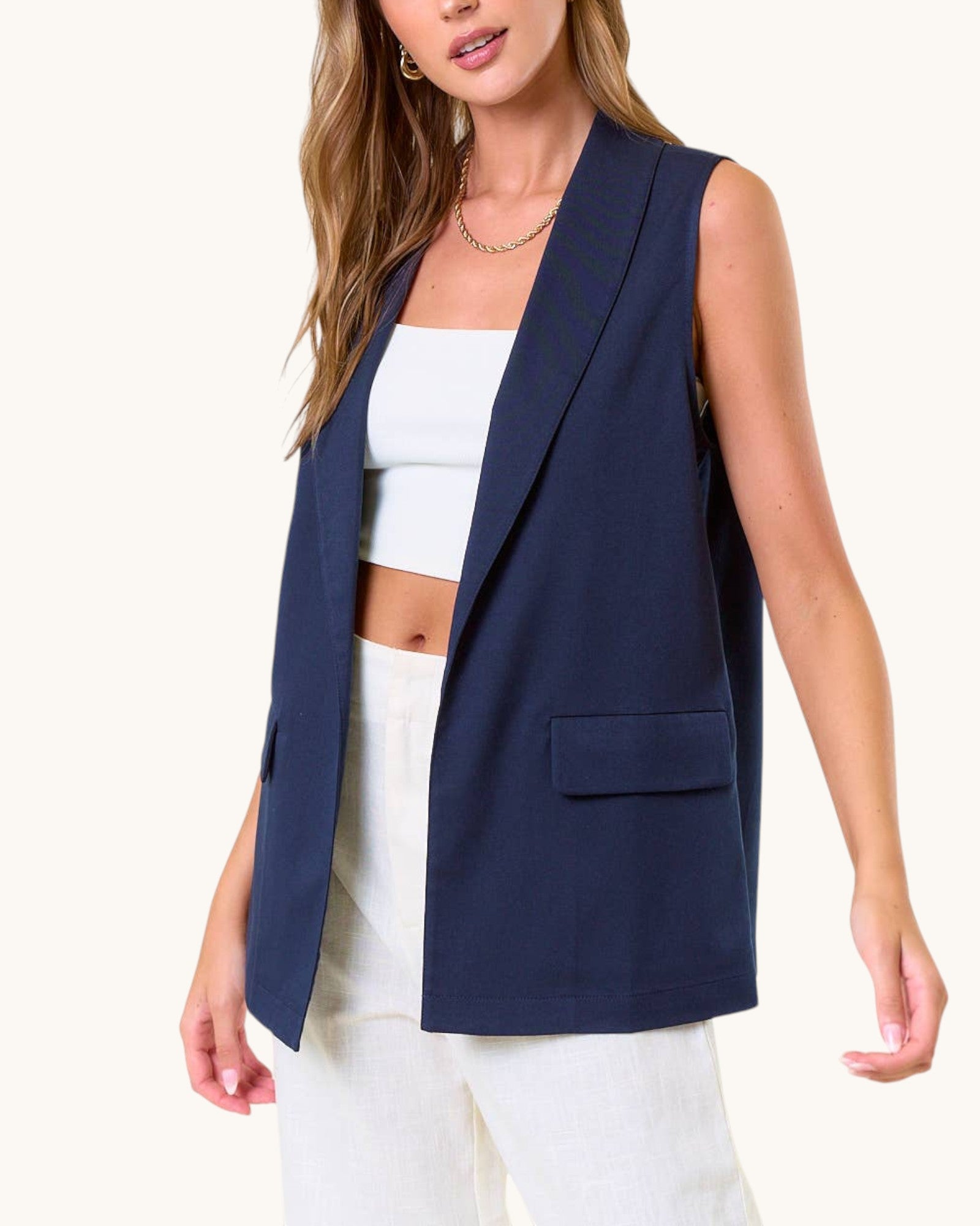 Woman wearing a navy blue sleeveless blazer over a white outfit on a white background.