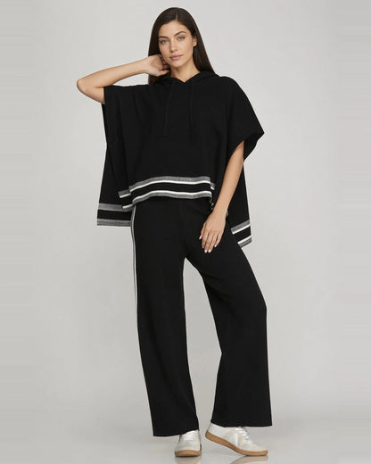 Full-length front view of model wearing black short sleeve knit hoodie cape with contrast striped hem and matching pants. 