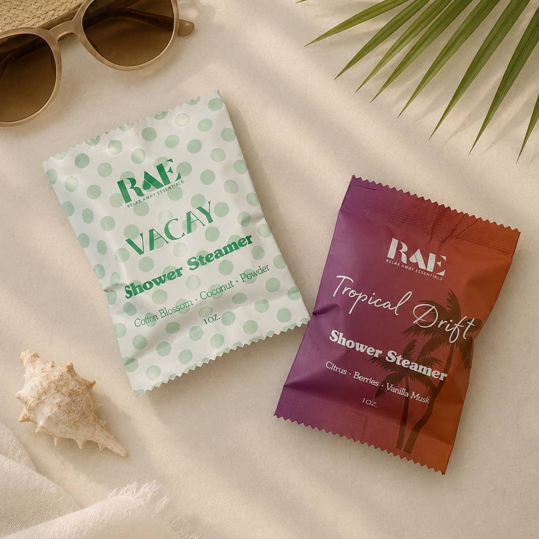 Two shower steamer packets from RAE on a sandy surface with sunglasses and a shell.