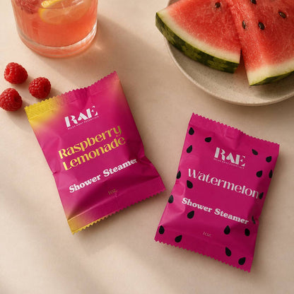 Two pink shower steamer packets labeled 'Raspberry Lemonade' and 'Watermelon' on a beige surface with watermelon slices and raspberries.
