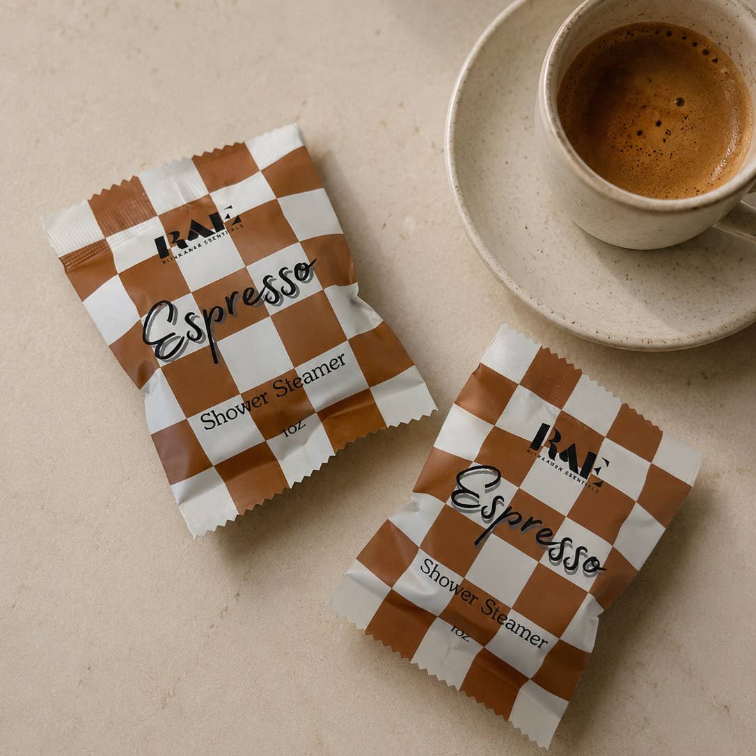 Two packets of Rae Espresso Shower Steamers on a beige surface with a cup of coffee.