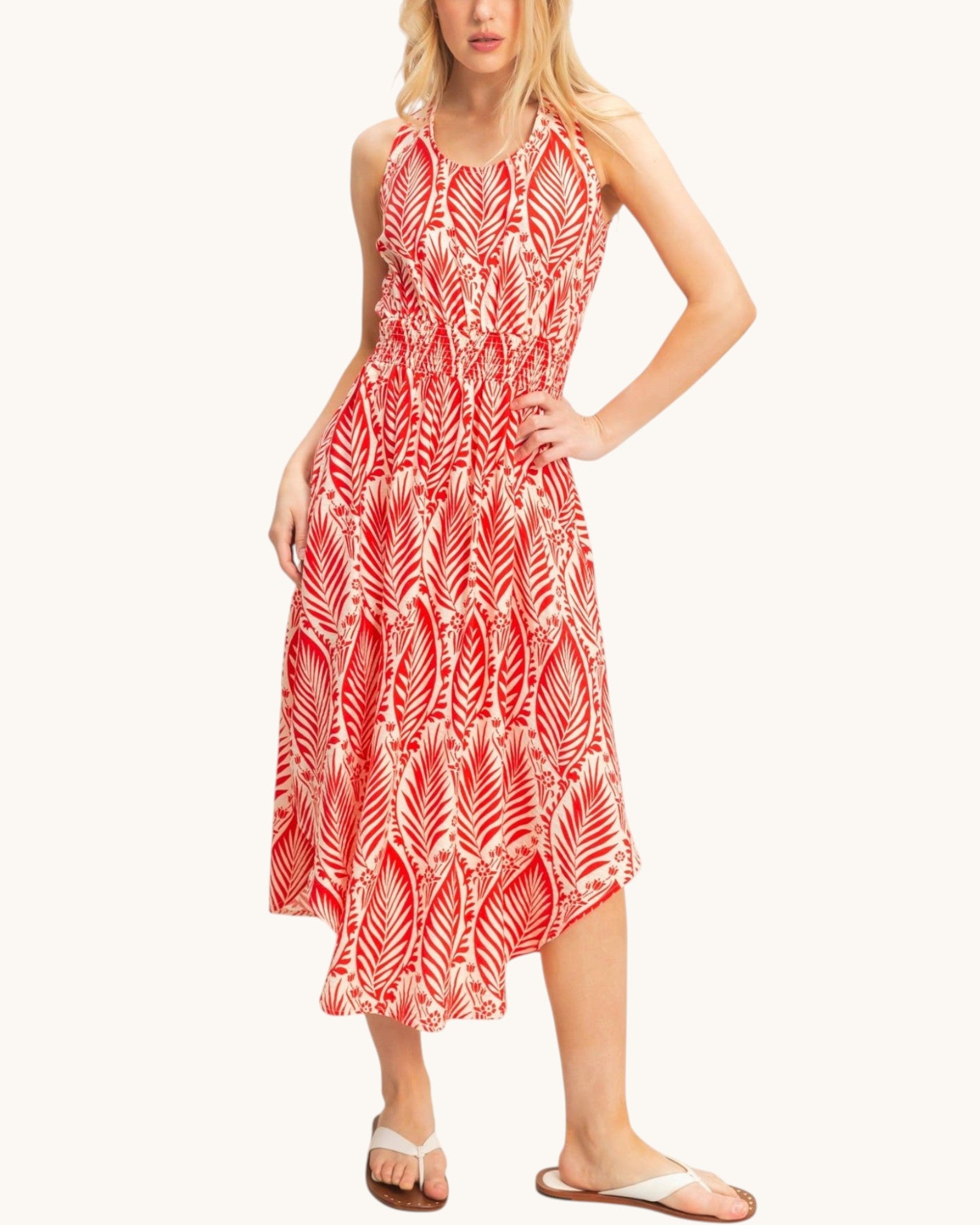 A woman modeling a sleeveless midi dress with a red and white palm leaf pattern, paired with white slide sandals, against a solid off-white background.