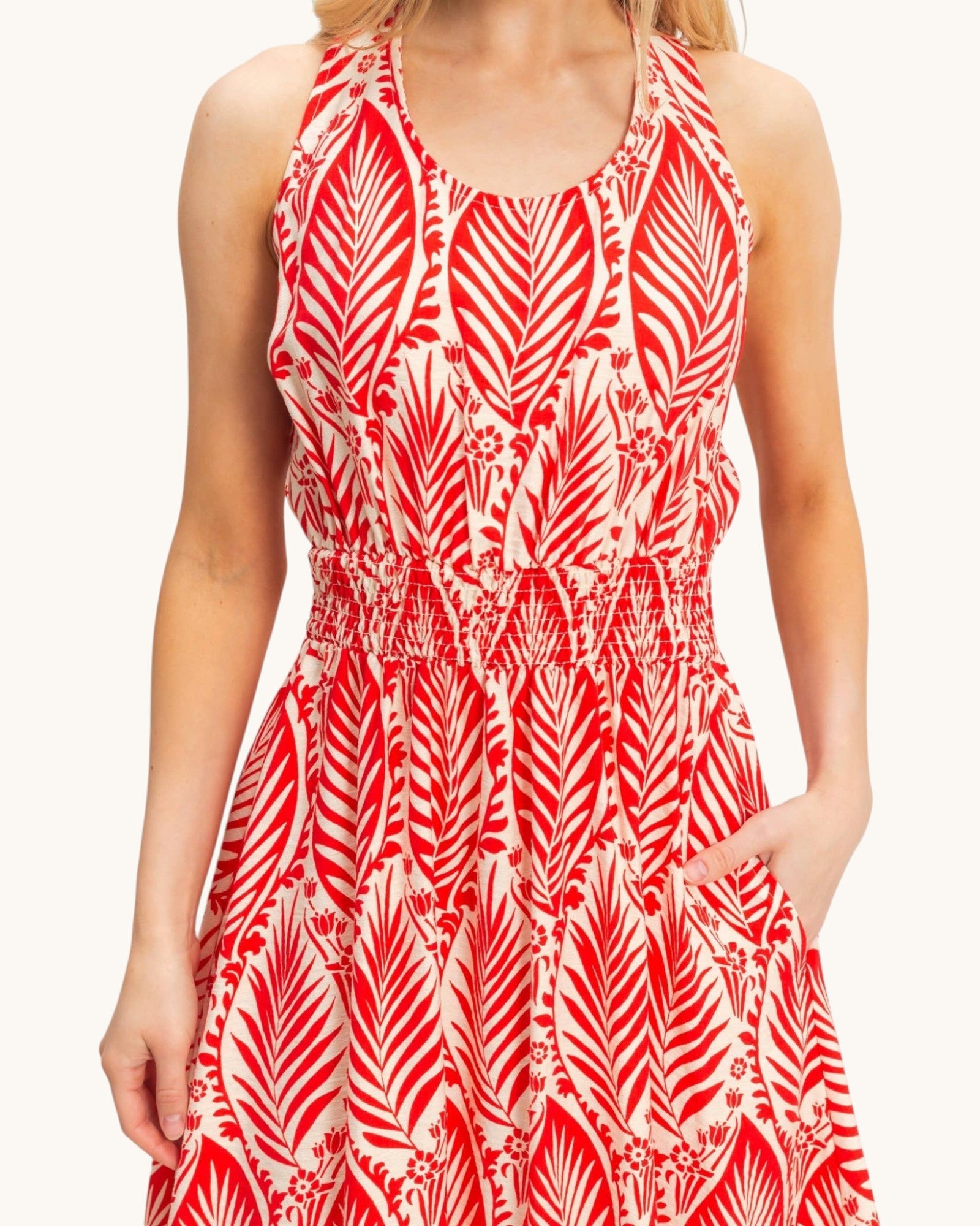 A sleeveless halter-neck dress with a red and white palm leaf pattern, featuring a smocked waist and side pockets, shown on a model against a light background.