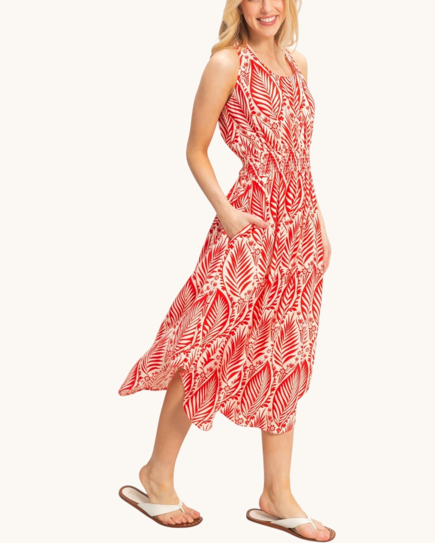 A woman modeling a sleeveless midi dress with a red and white palm leaf pattern, featuring side pockets and a smocked waist, paired with white slide sandals against a solid off-white background.